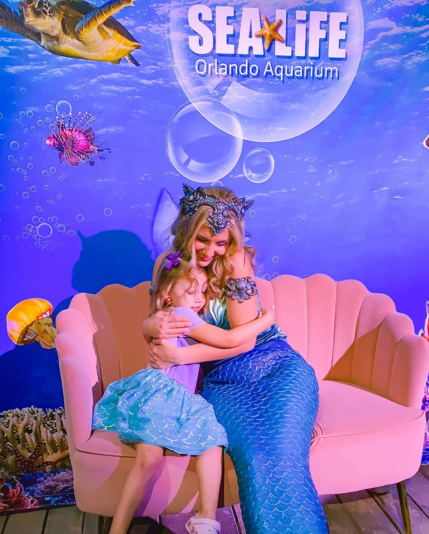 A mermaid embraces a young girl on a pink sofa at the SEA LIFE Orlando aquarium, with ocean-themed decor and logo in the background.